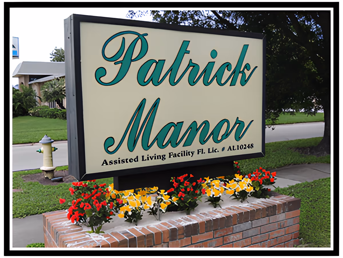 Outdoor sign reading "Patrick Manor" mounted above a brick planter with red and yellow flowers in front of the facility.