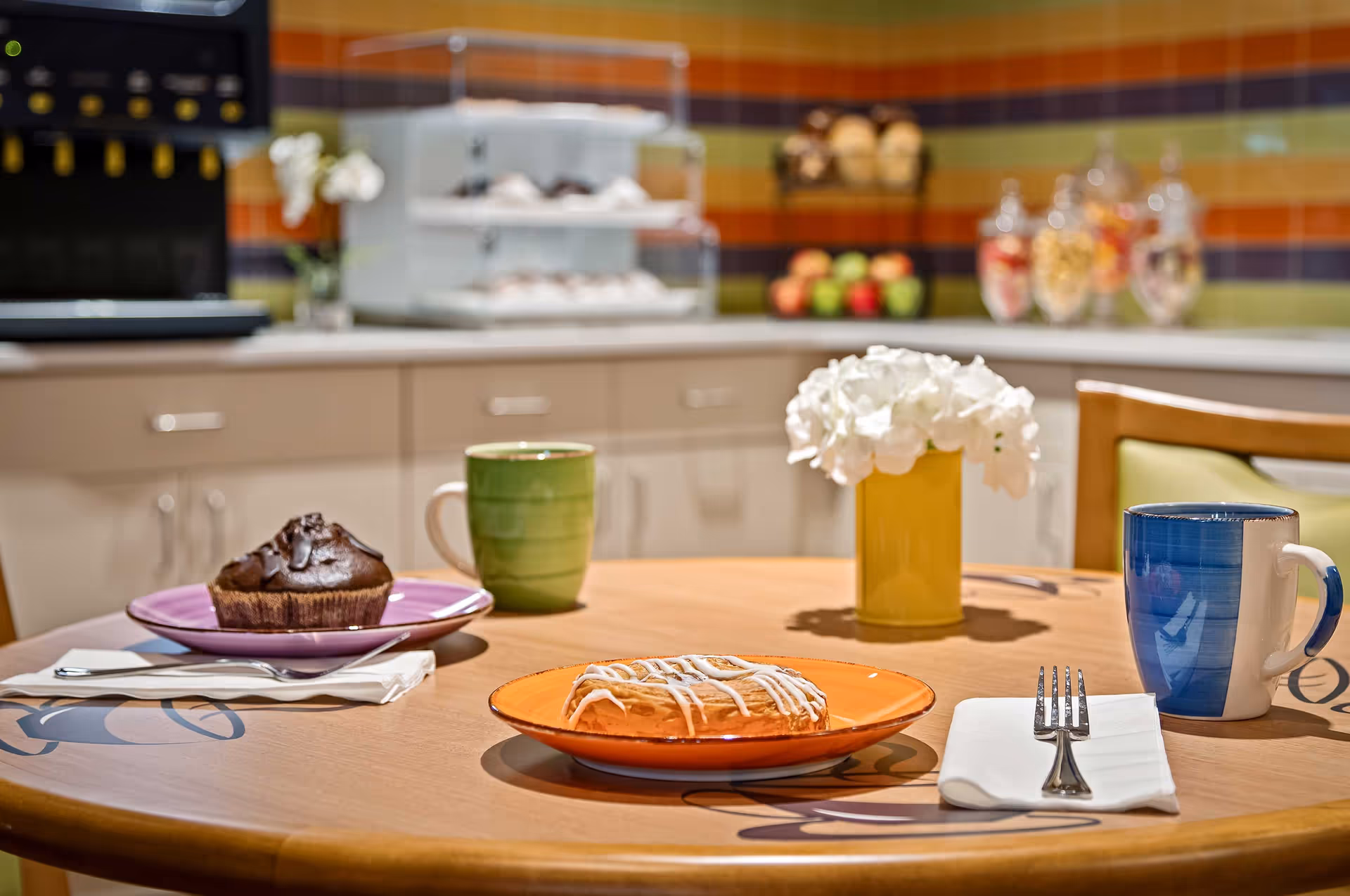 A wooden table set with a chocolate muffin on a purple plate, a pastry with white icing on an orange plate, a green mug, a blue and white mug, a fork on a white napkin, and a yellow vase with white flowers. In the background, there is a kitchen counter with a coffee machine, a display case with pastries, and jars of snacks against a colorful tiled wall.