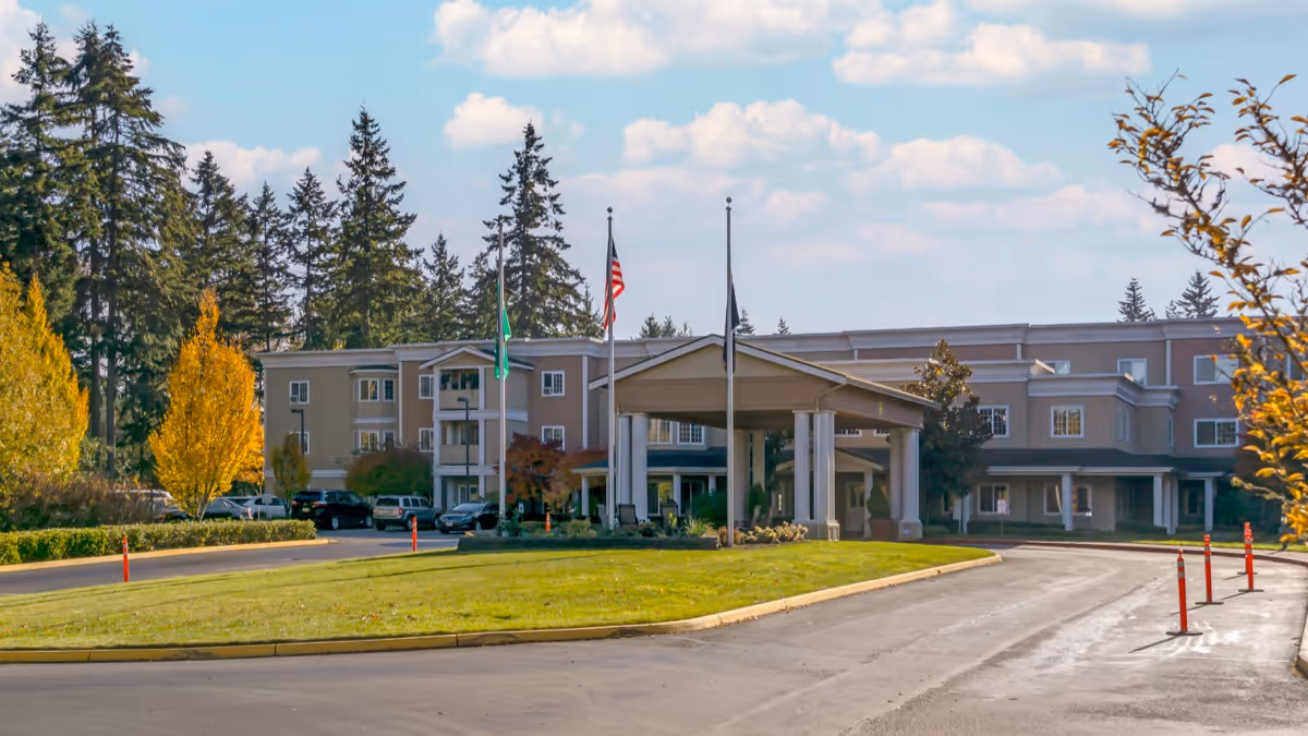 Exterior view of Brookdale Foundation House, a multi-story senior living facility with a covered entrance, surrounded by trees and a well-maintained lawn under a partly cloudy sky.