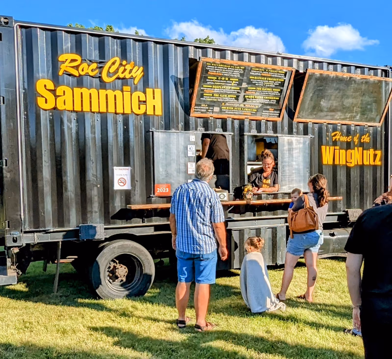 A black mobile food truck named Roc City Sammich serves customers outside on grass.