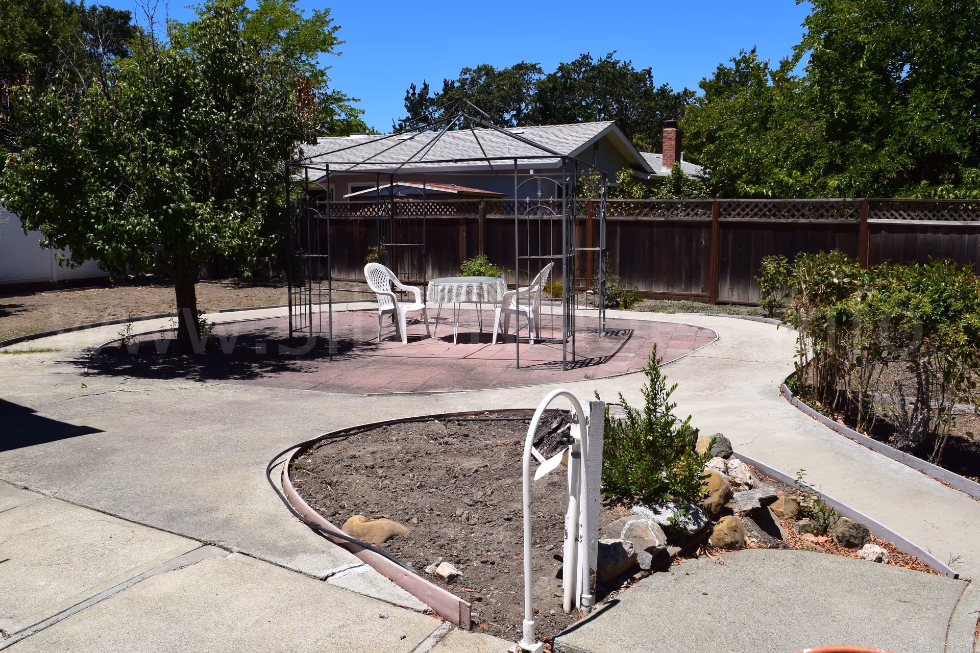 Outdoor courtyard featuring a decorative metal gazebo, plastic chairs around a table, with surrounding concrete paths.