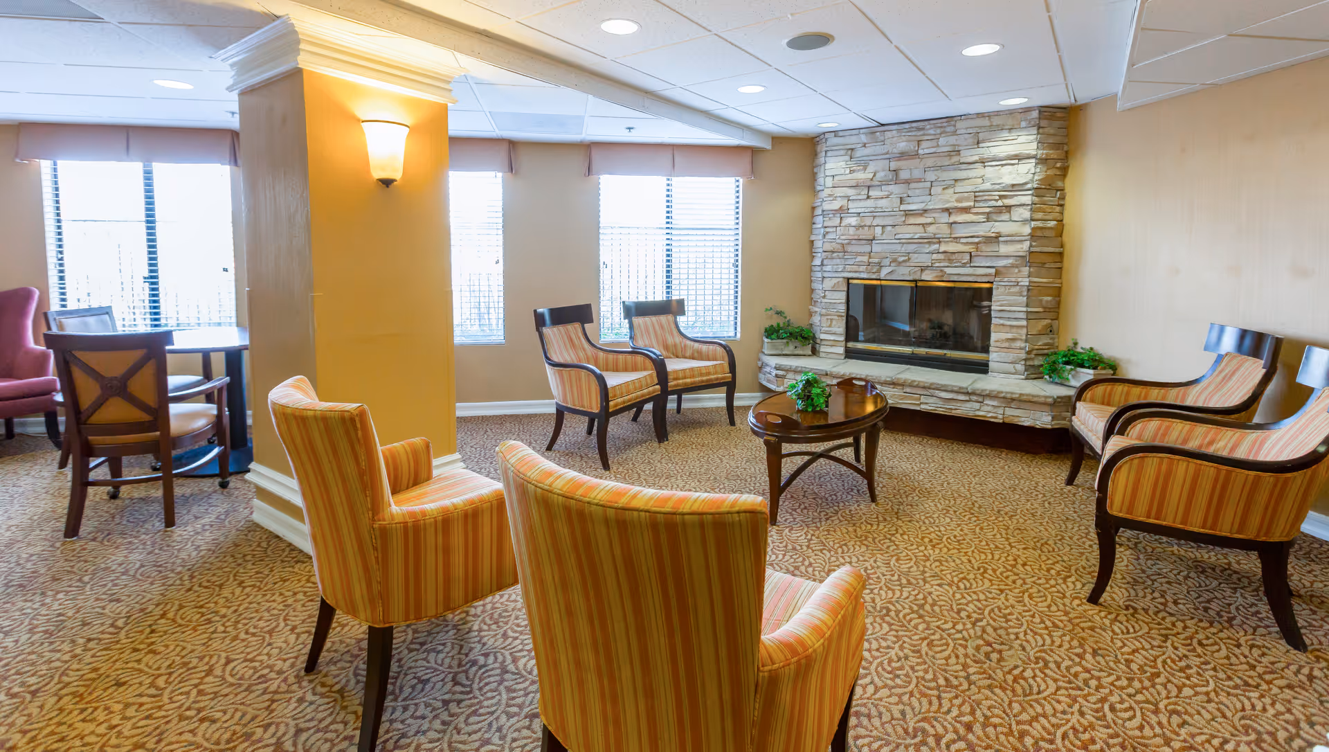 Cozy senior living lounge with striped chairs, coffee table, fireplace, and large windows.