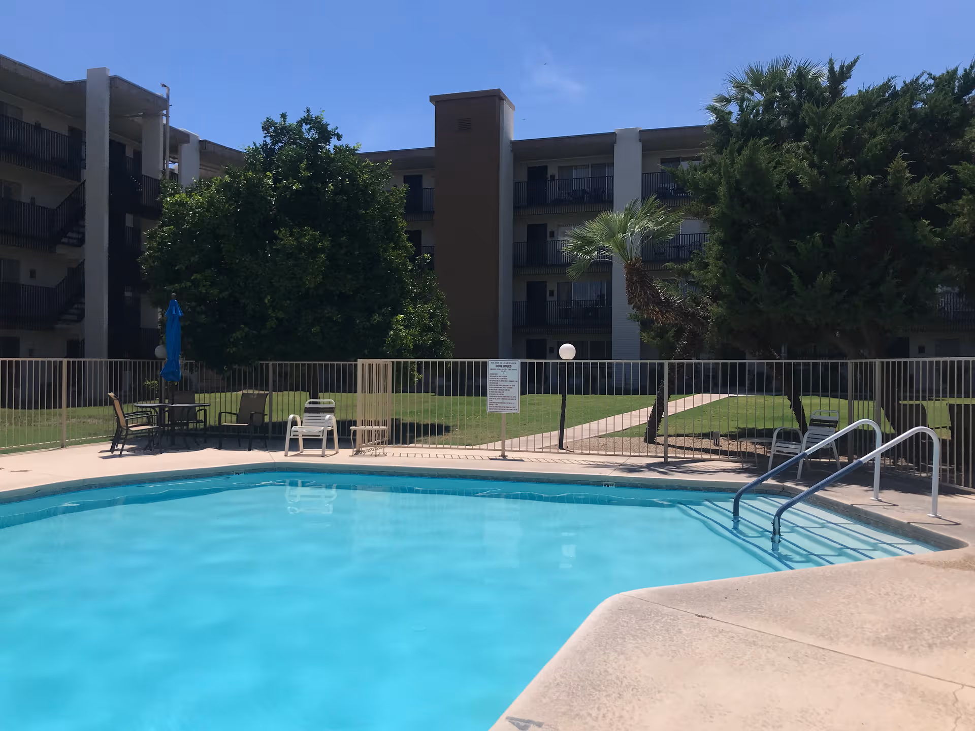 Outdoor pool area with lounge chairs, umbrella table, and multi-story building behind.