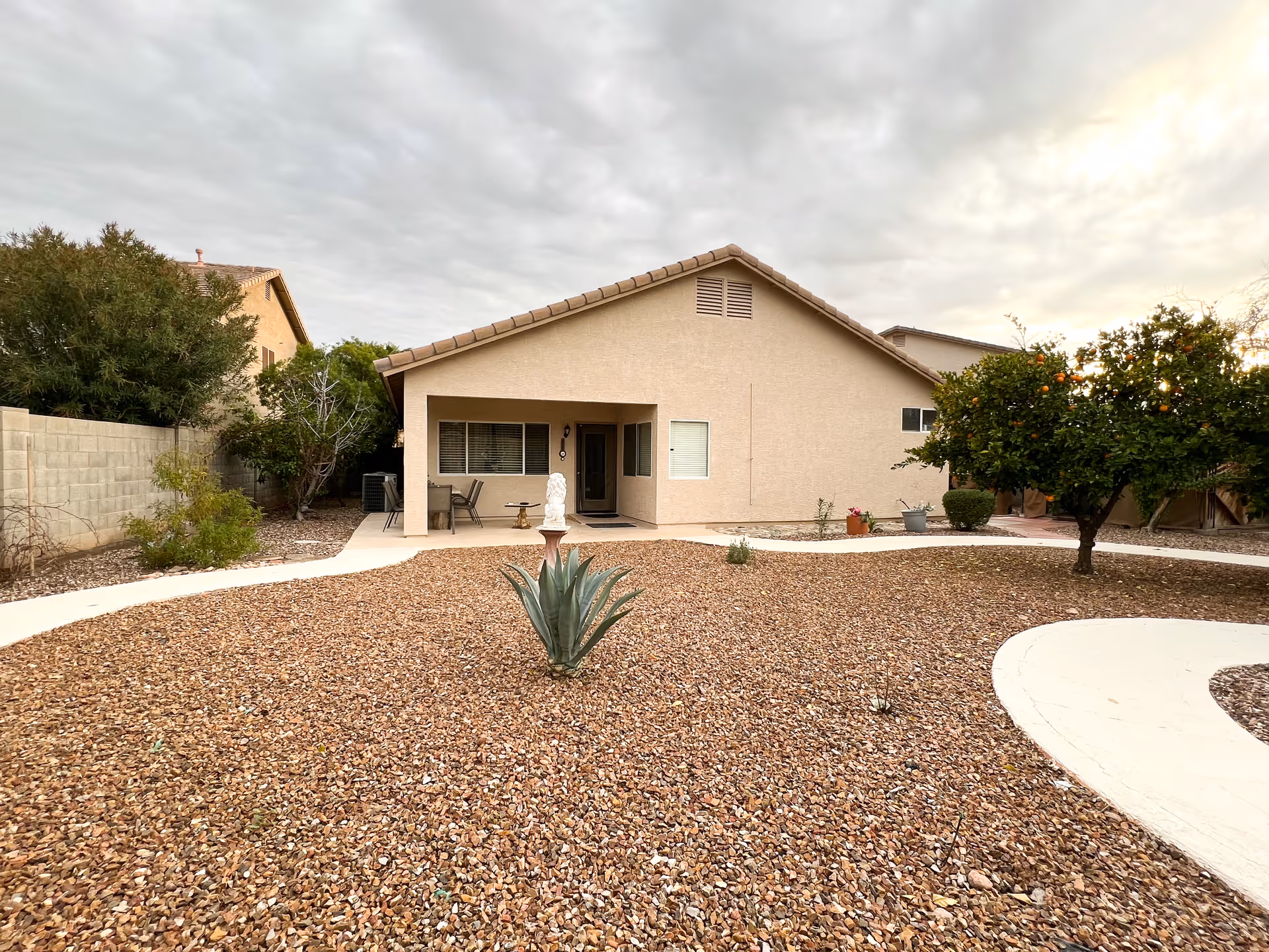 Single-story beige house with a covered patio, gravel yard with an agave plant and an orange tree under a cloudy sky.