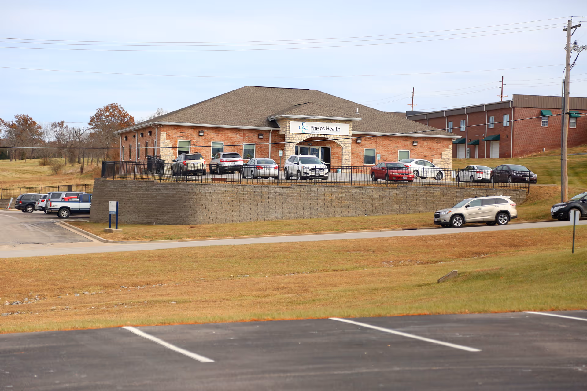 Exterior view of a brick Phelps Health building with a parking lot and cars.