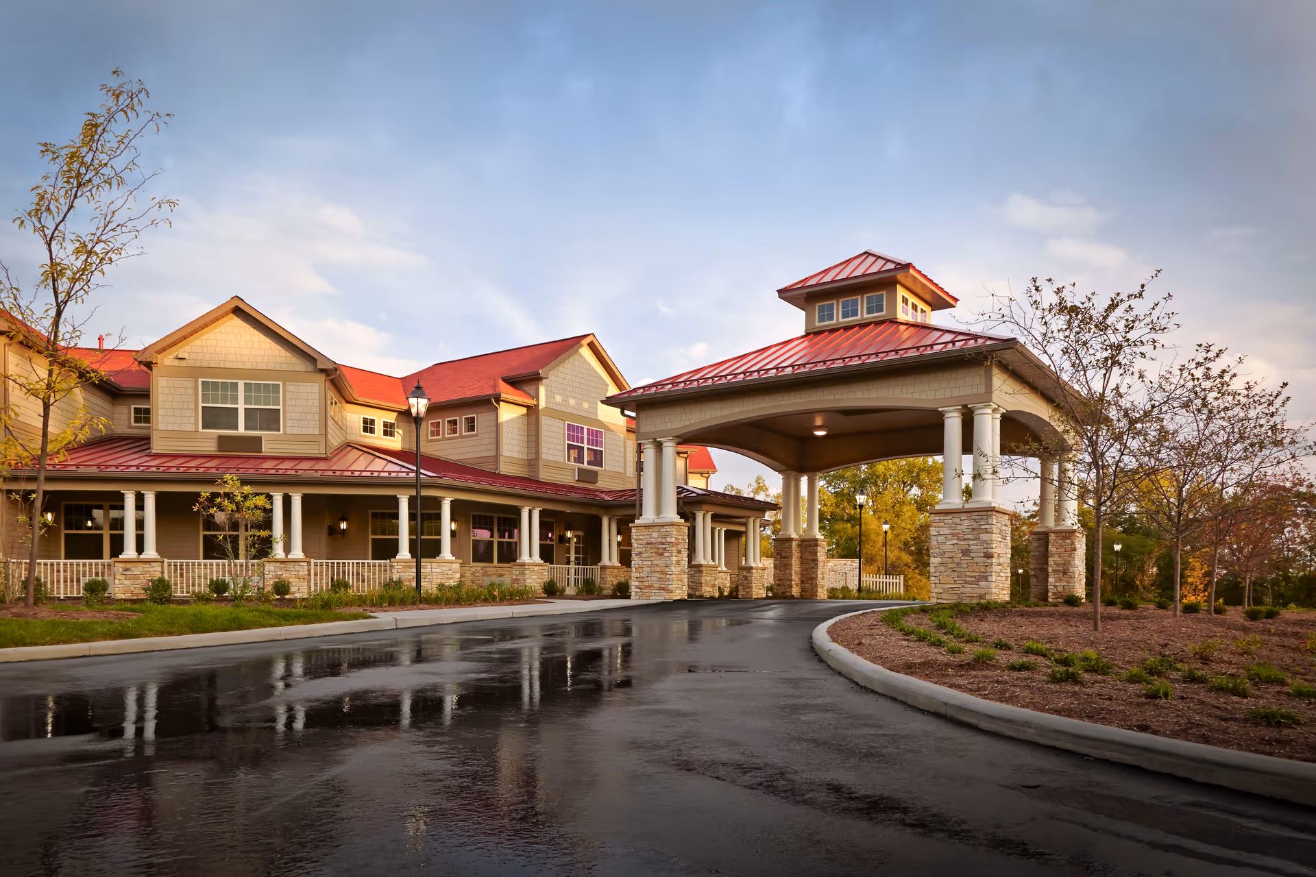 Exterior view of a senior living facility with a covered entrance supported by stone pillars and white columns. The building has beige siding with red roofs and multiple windows. The driveway is wet, reflecting the building and surrounding trees with autumn foliage under a partly cloudy sky.