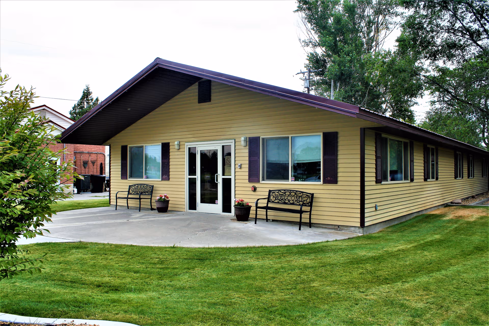 Front view of a single-story beige assisted living building with benches and a grassy lawn.
