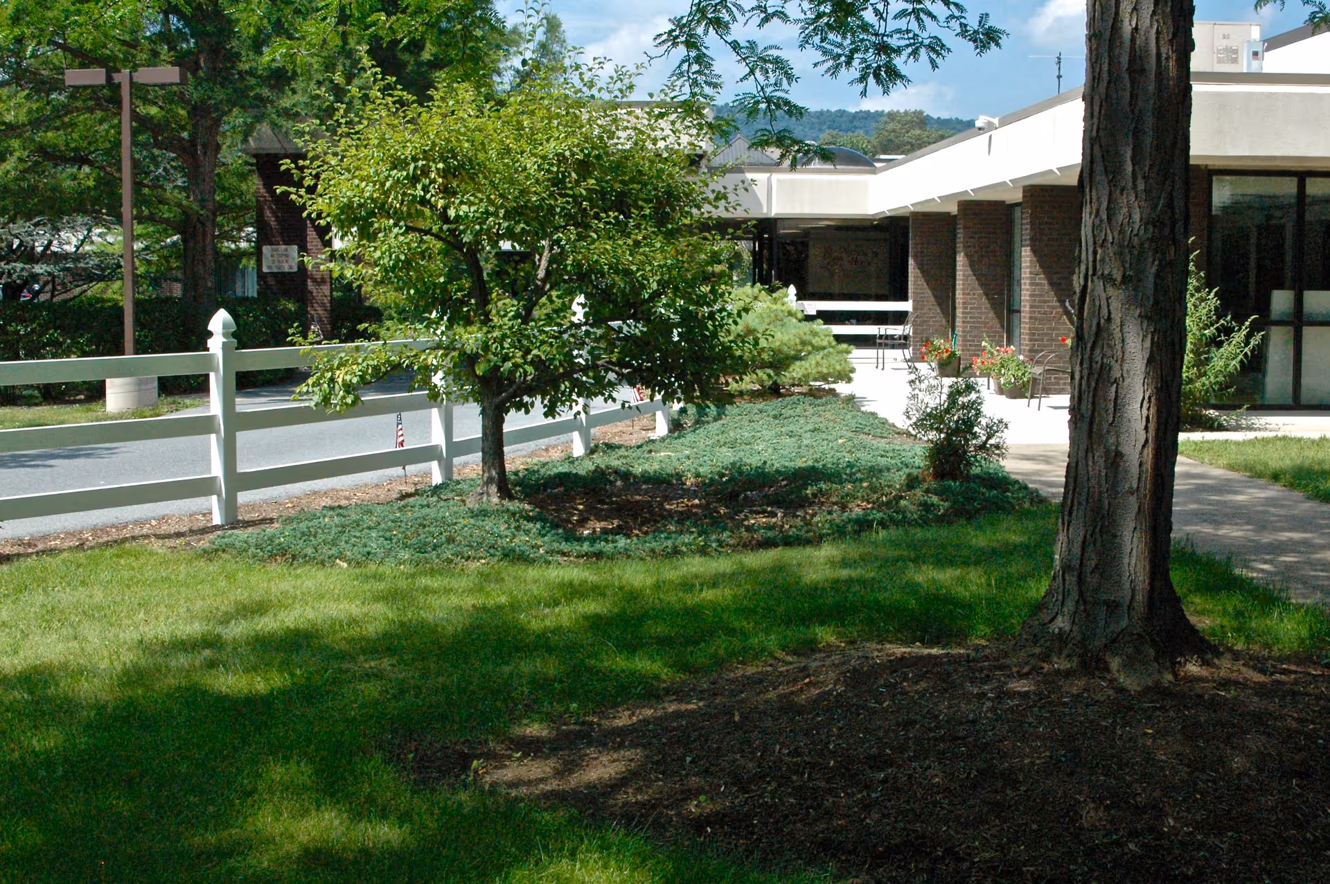 Front exterior of a senior living facility with a grassy lawn, trees, a white fence, and the building entrance with planters.