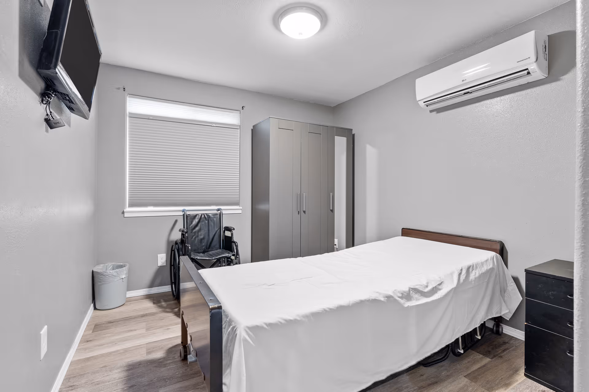 A compact senior bedroom with a single bed, wheelchair, wardrobe, TV, and wall-mounted air conditioner.