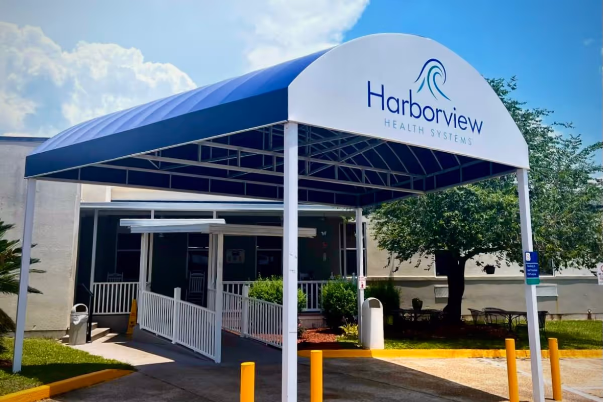 Entrance to Harborview Health Systems with a blue and white canopy over the walkway leading to the building entrance. There are yellow bollards, a trash can, and some greenery including a tree and bushes near the entrance.