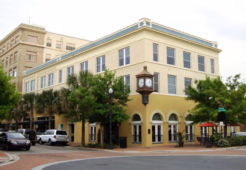 Two-story beige building with arched entrances, street clock, trees, and outdoor seating along the sidewalk.