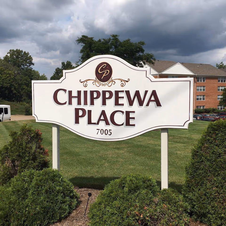 A large white sign with decorative edges displaying the text 'Chippewa Place 7005' in maroon letters, set on a green lawn with bushes in the foreground and a multi-story brick building in the background under a cloudy sky.