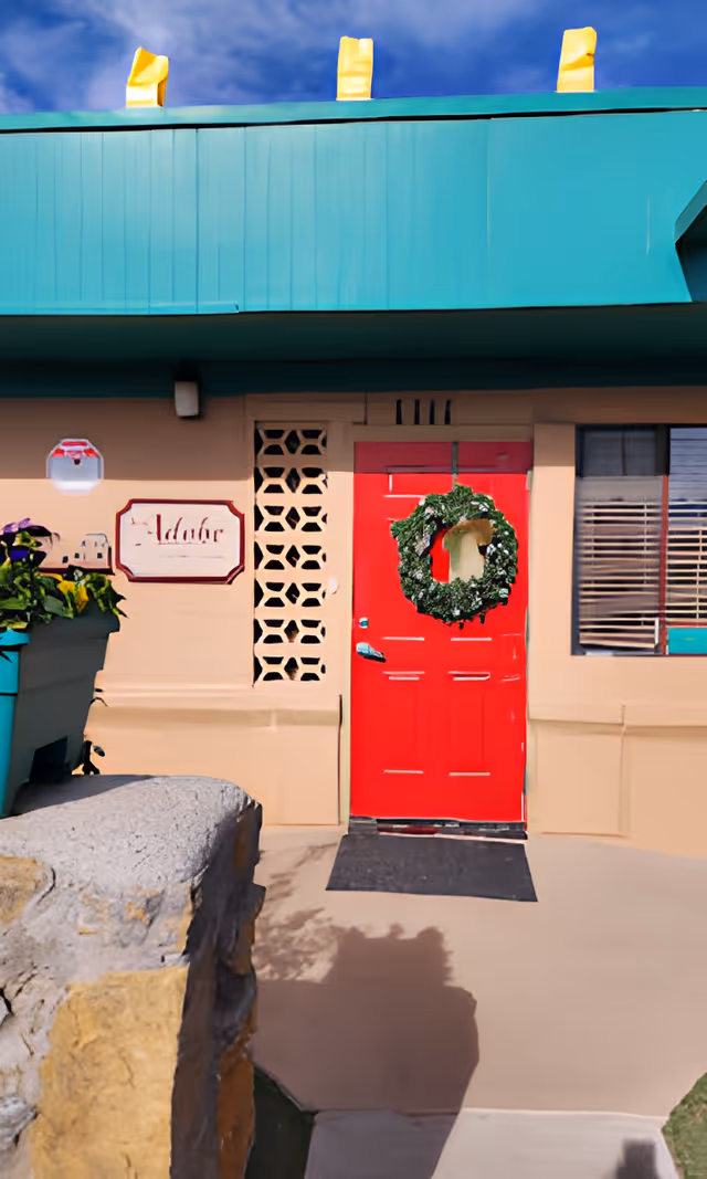 Exterior view of a building entrance with a bright red door decorated with a green wreath. The building has beige walls, a teal roof, and a sign next to the door that reads 'Adobe'. There is a window with blinds to the right of the door and some plants in a planter to the left.