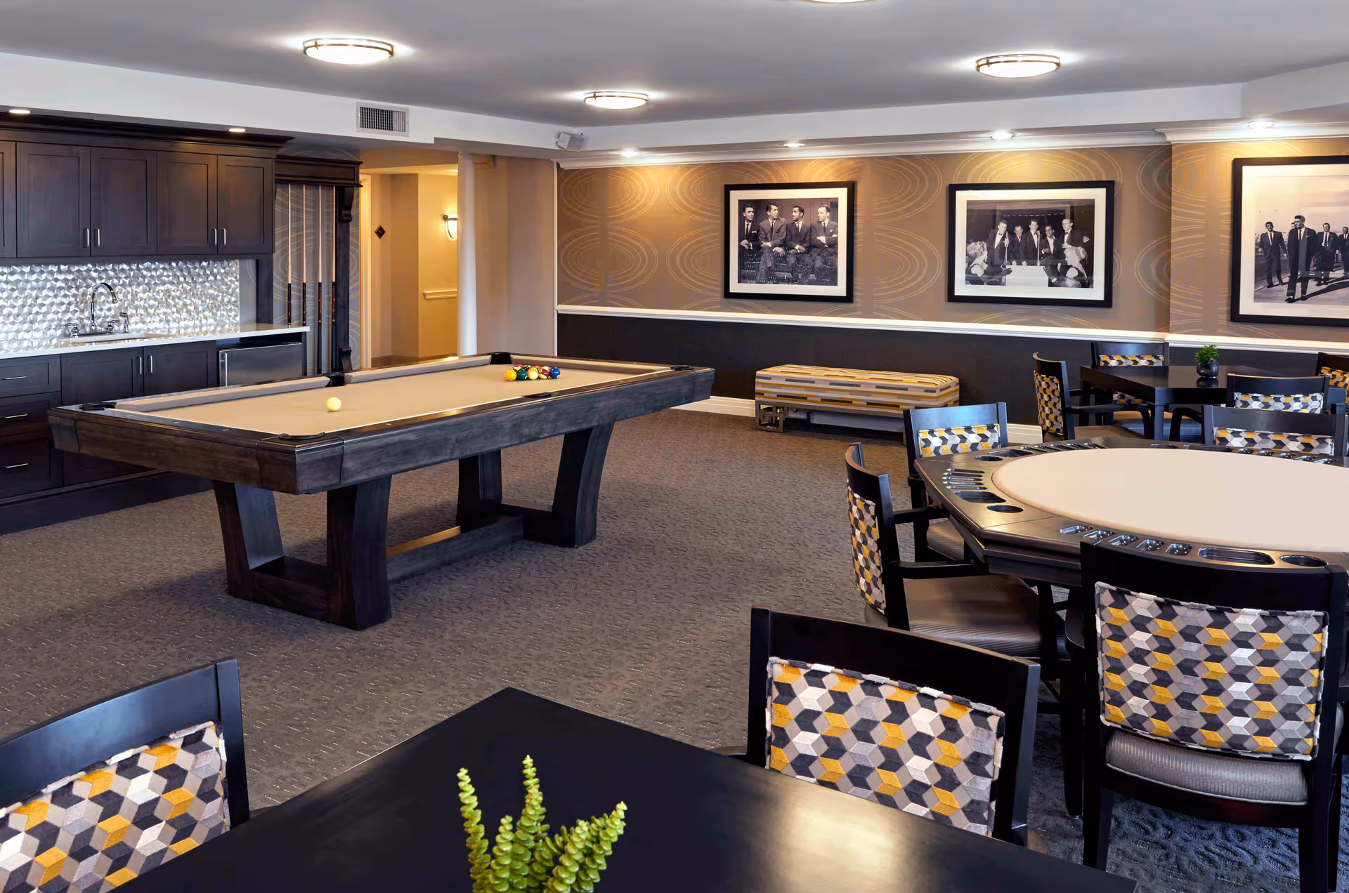 A spacious recreational room with a pool table in the center, surrounded by tables and chairs with patterned upholstery. The room features dark cabinetry with a sink and a textured backsplash on one side. The walls are decorated with framed black and white photographs, and the lighting is provided by ceiling-mounted fixtures.