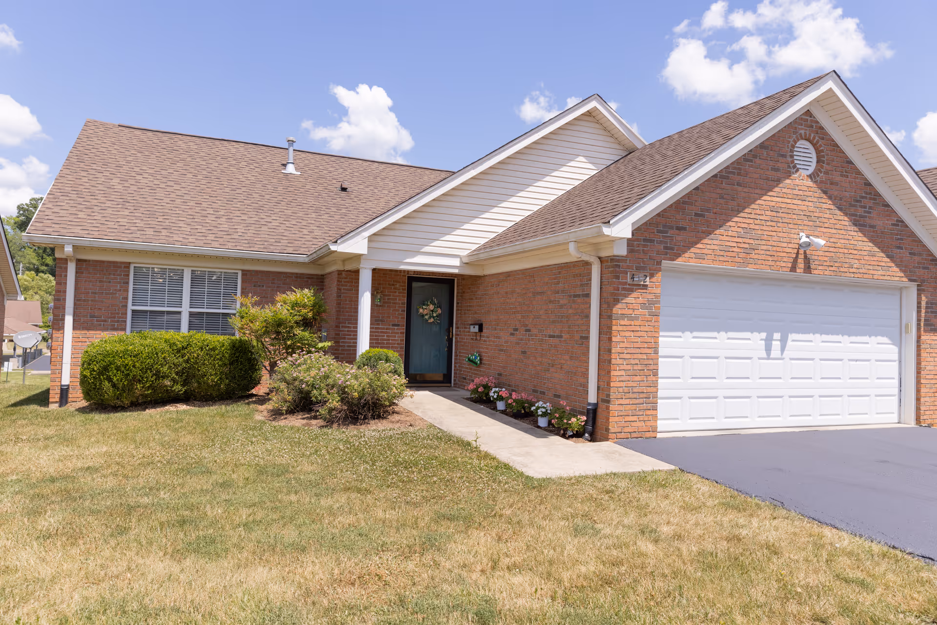 Front view of a single-story brick house with a white two-car garage, blue entry door with a wreath, and a grassy front yard.