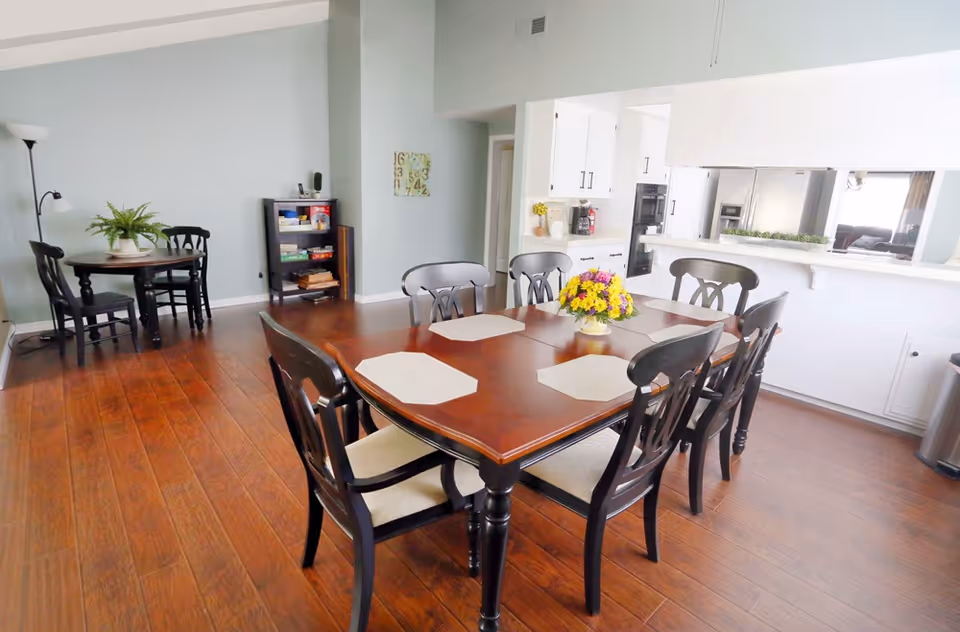 Spacious dining area with a wooden table set for six, a floral centerpiece, and a view into a white kitchen and small sitting area.