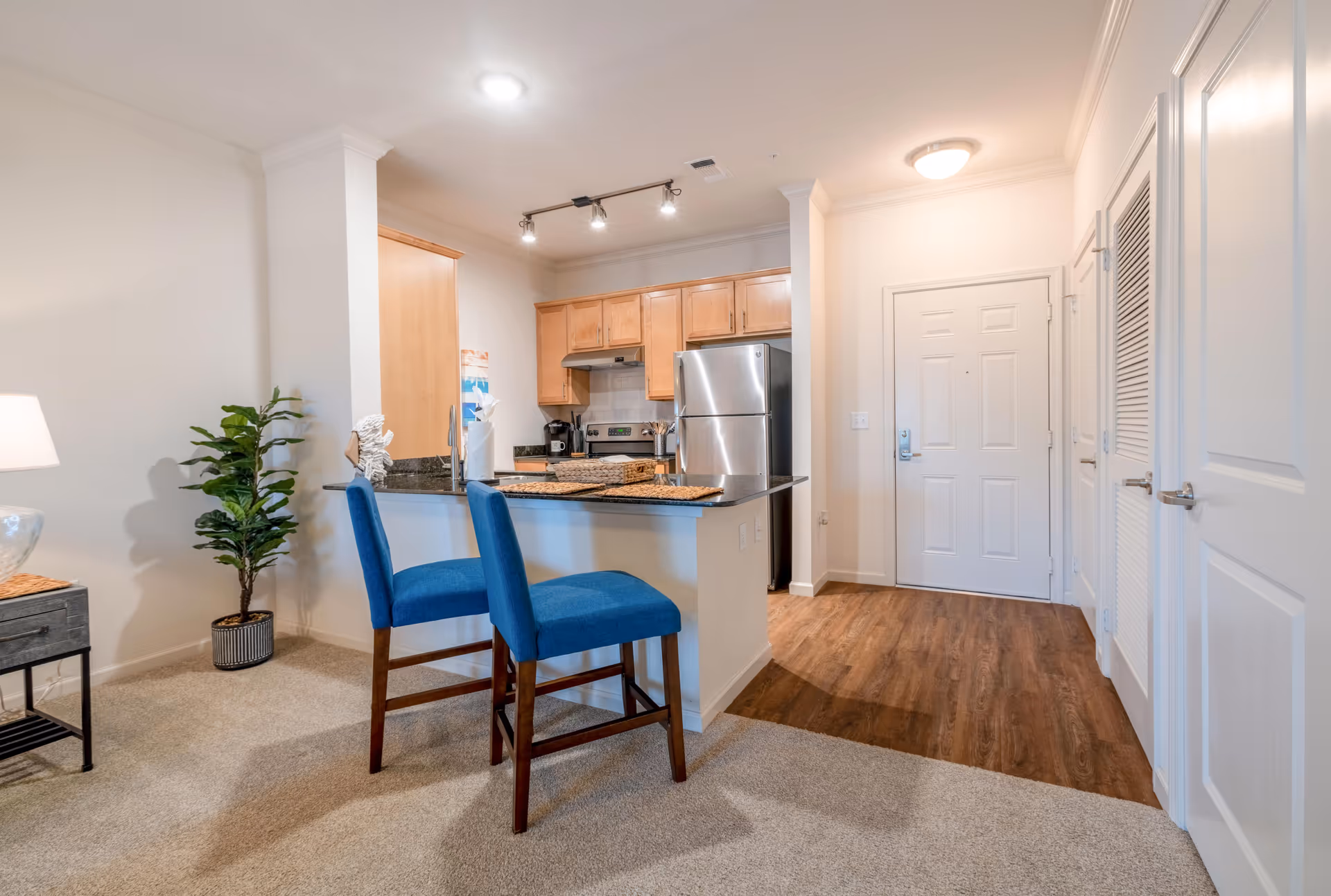 Open-concept kitchenette with wood cabinets, stainless appliances, granite counters, and blue bar chairs.