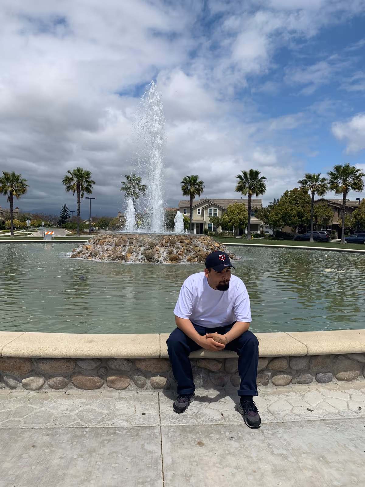 Person sits on fountain wall in an outdoor plaza with palm trees.