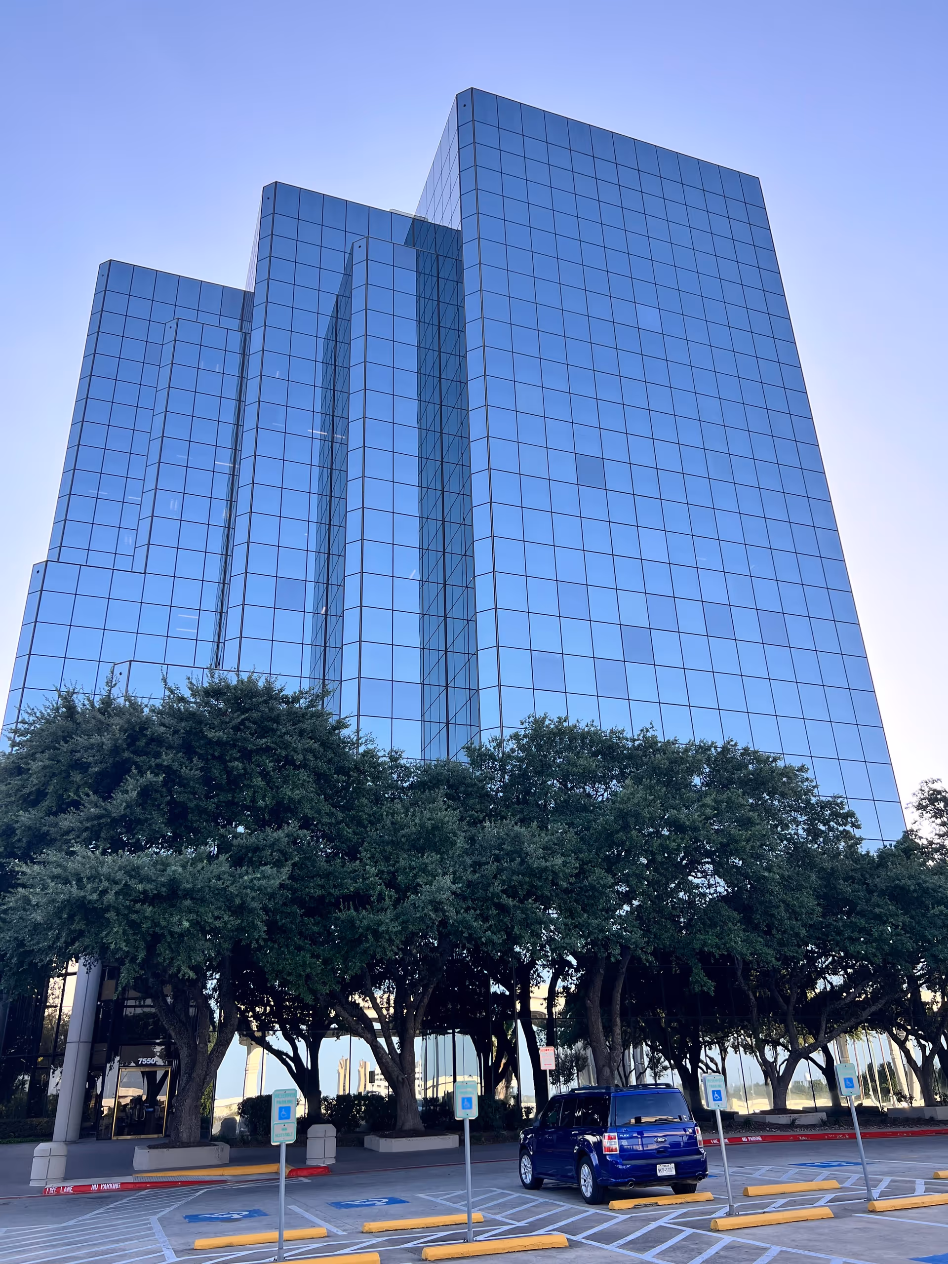 Tall glass office building with trees in foreground and a blue SUV in a parking lot.
