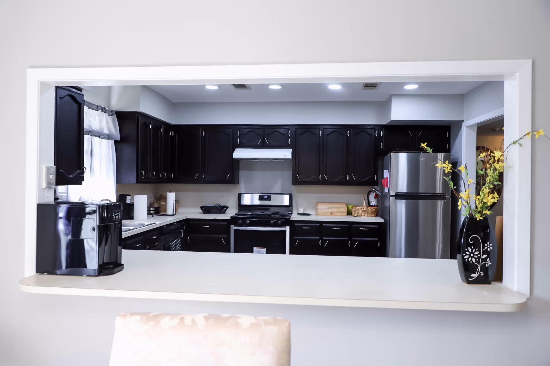 View through a pass-through window into a kitchen with dark wood cabinets, a stainless steel refrigerator, a stove with a white range hood, and a coffee maker on the counter. A black vase with yellow flowers is placed on the counter in the foreground.