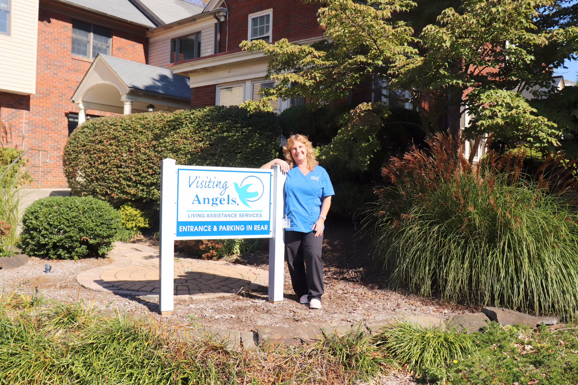 Caregiver in blue scrubs stands beside Visiting Angels sign at the senior home's landscaped front.