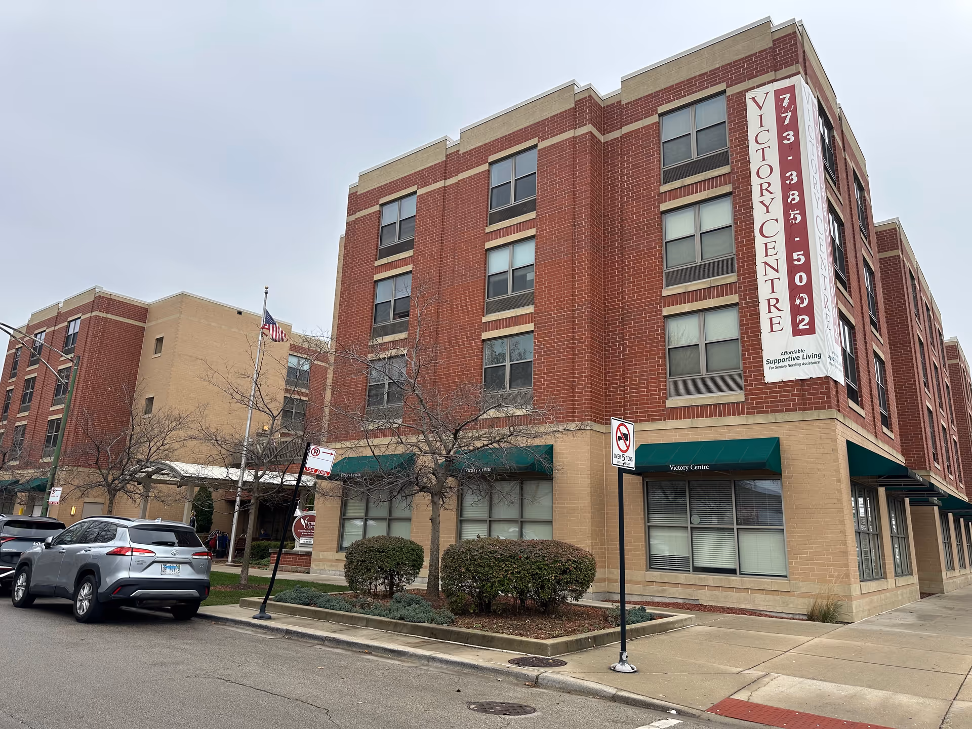 Exterior view of a multi-story brick and beige building with green awnings labeled Victory Centre. There are leafless trees and parked cars along the street in front of the building. A large vertical banner on the corner of the building displays the name Victory Centre and a phone number.
