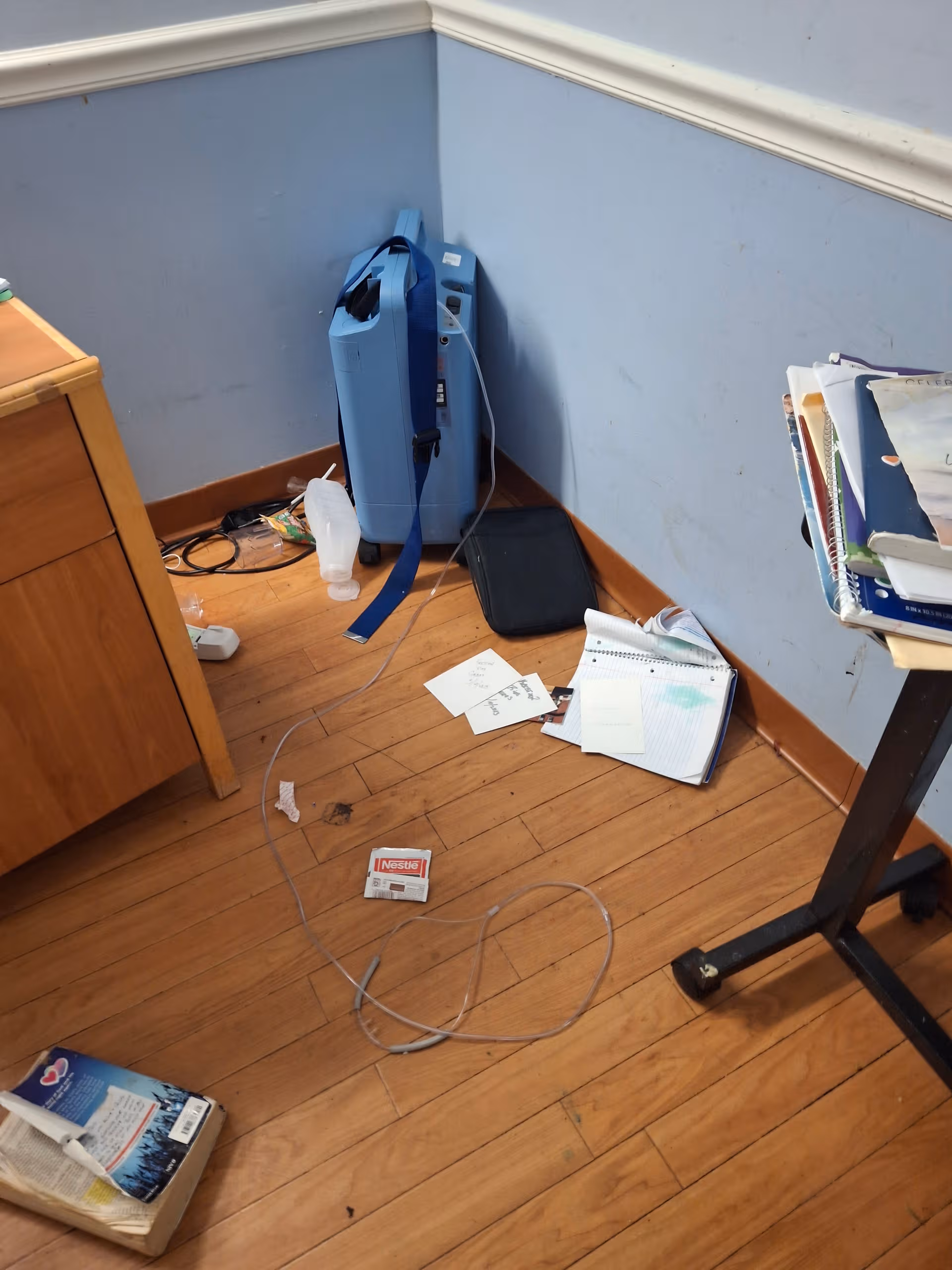 Corner of a cluttered room with a blue medical device, papers, cables, and a rolling desk.