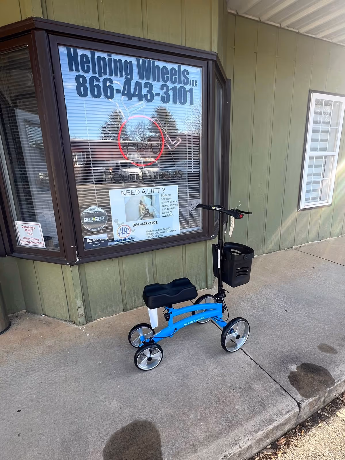 Blue mobility scooter parked outside a Helping Wheels Inc storefront window.