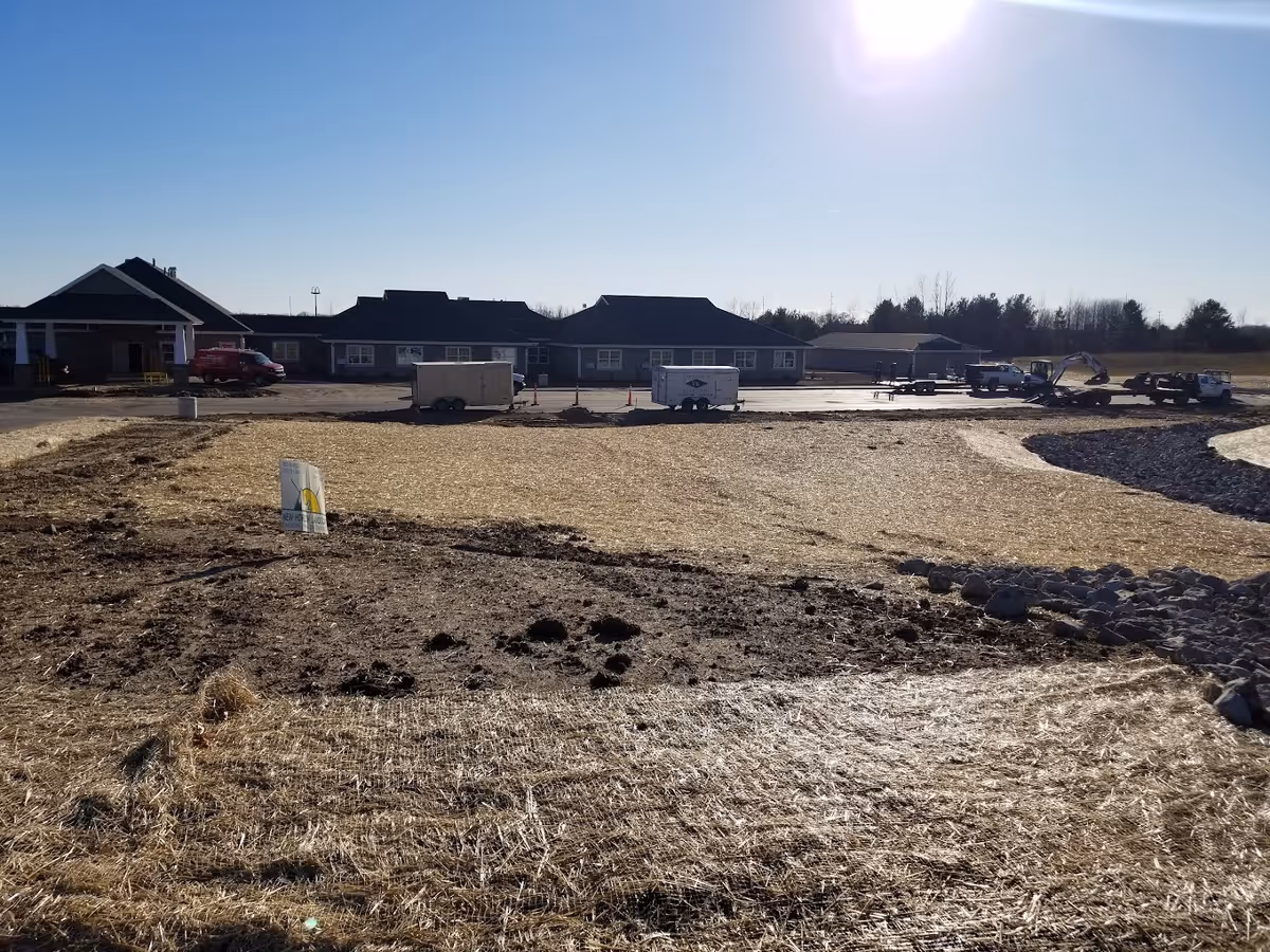 Front view of a single-story senior living building and surrounding construction site with trailers, equipment, and straw-covered ground under a bright sun.
