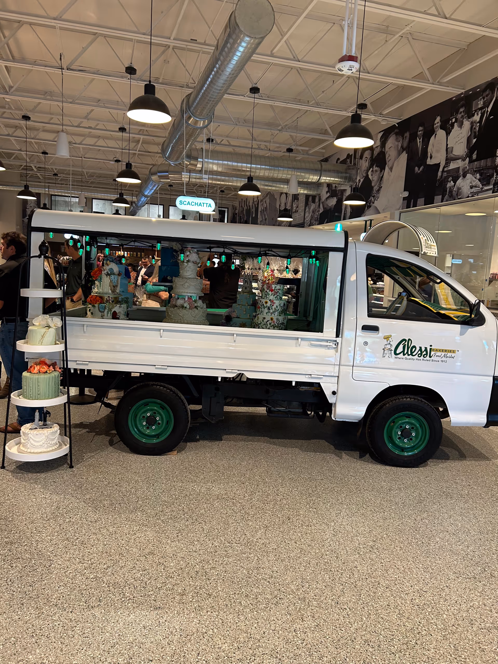 White delivery van with green wheels parked inside a showroom beside multi-tiered cakes on display.