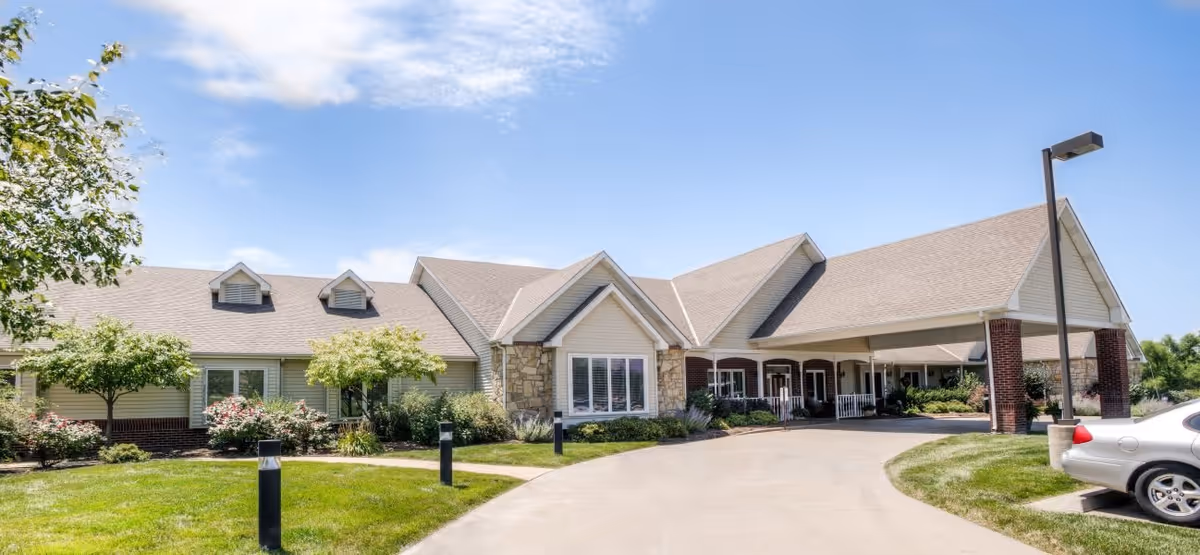 Exterior view of a single-story senior living facility building with a covered entrance driveway, surrounded by green lawns, small trees, and shrubs under a partly cloudy blue sky.
