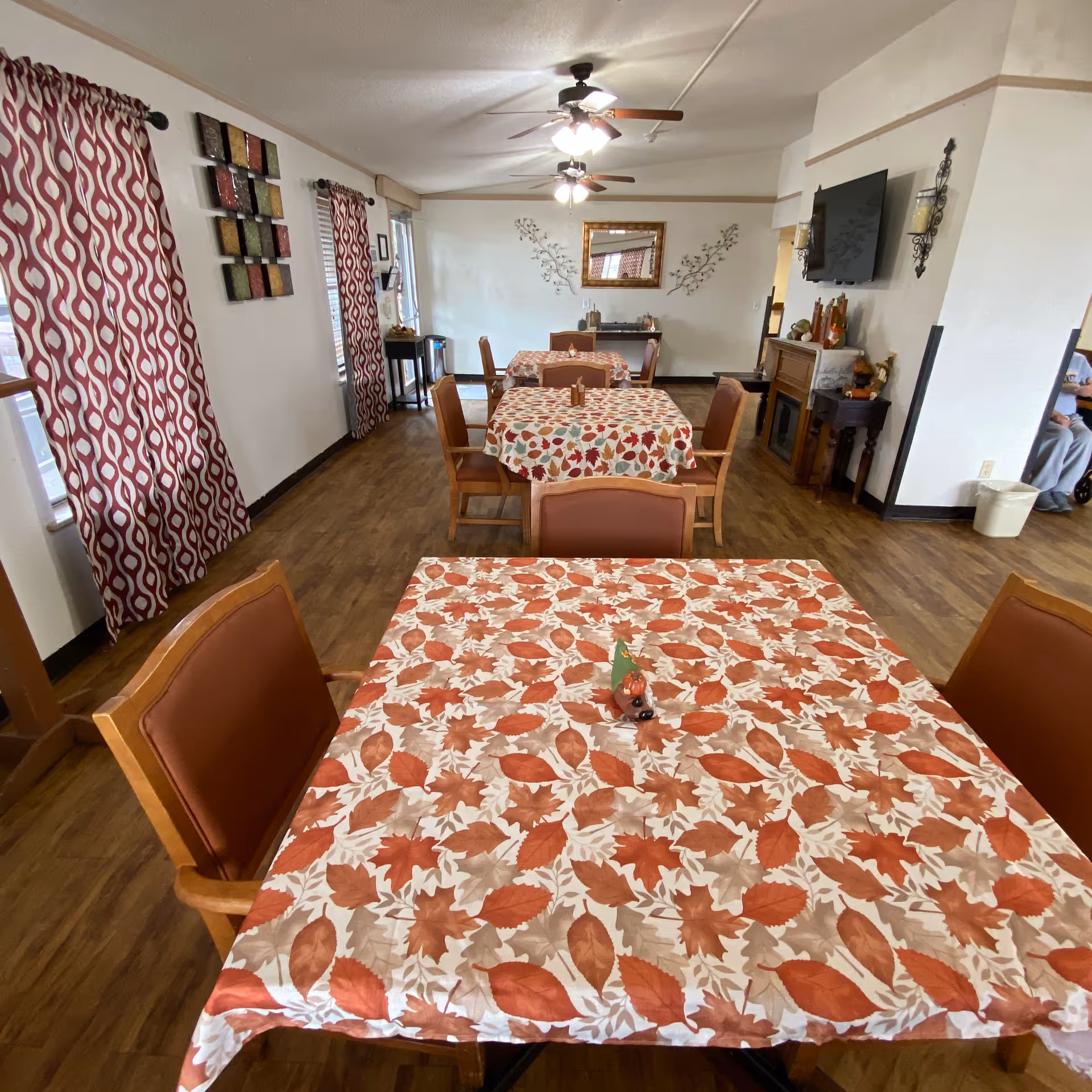 A dining room with several tables covered in autumn-themed tablecloths featuring orange and beige leaves. Each table is surrounded by wooden chairs with brown cushions. The room has wood flooring, patterned red and white curtains on the windows, ceiling fans with lights, and a wall-mounted TV above a fireplace. Decorative wall art and a mirror are visible on the far wall.