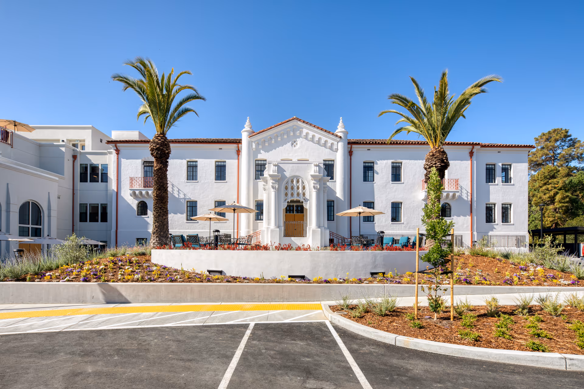 Front exterior of a white Mediterranean-style three-story building with palm trees, landscaped flower beds, and outdoor seating seen from the parking area.