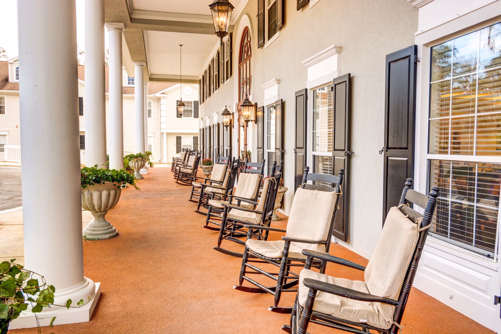 Covered front porch with a row of cushioned rocking chairs, large white columns, planters and hanging lanterns.