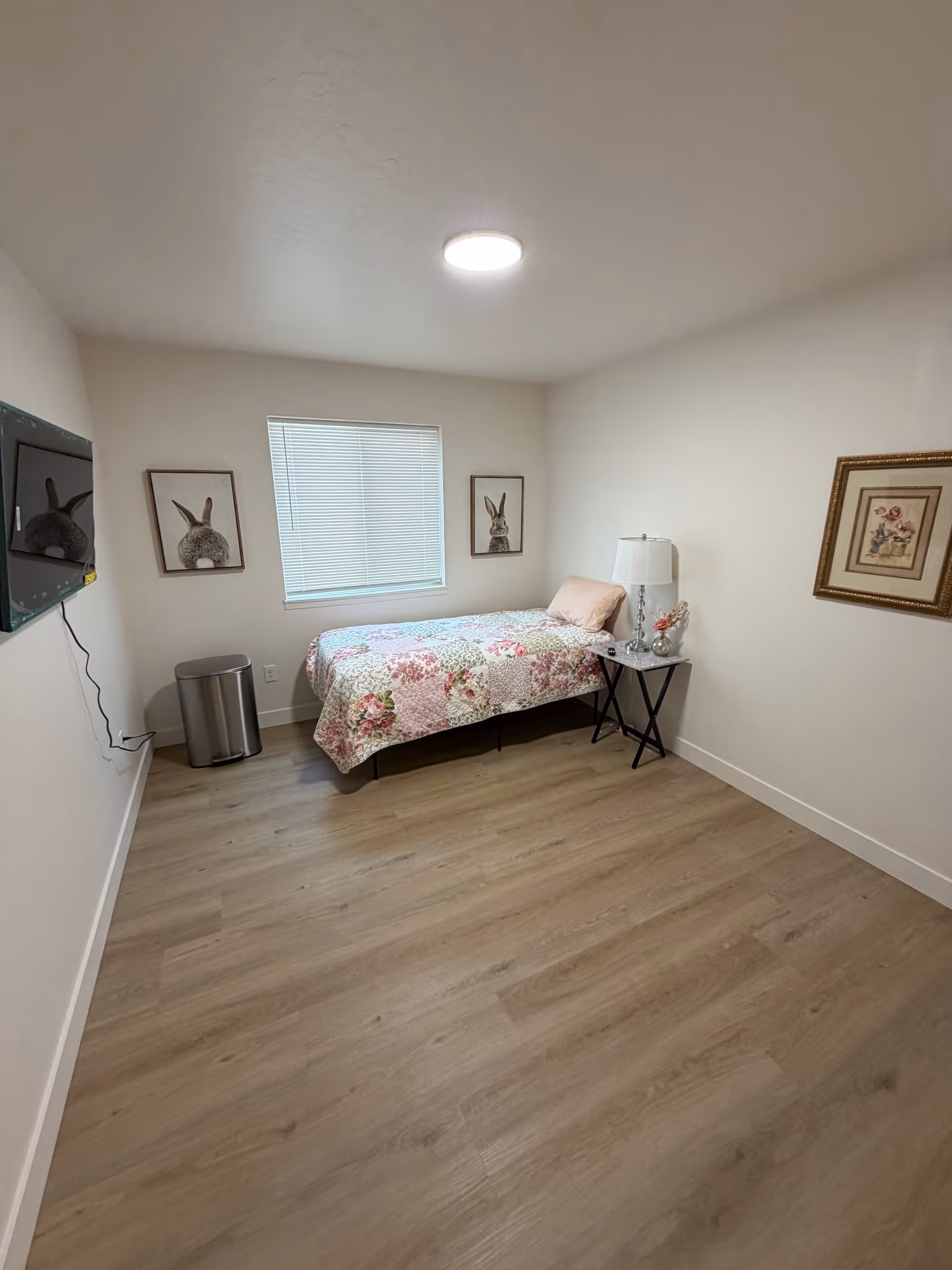Small bedroom with floral bedspread, nightstand, lamp, wall art, and mounted TV.