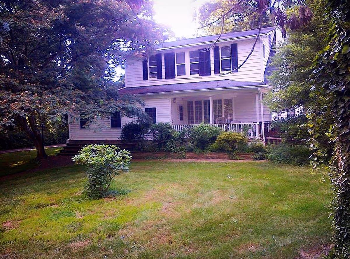 A two-story white house with black shutters surrounded by lush greenery and trees. The house has a covered porch with white railings and several windows. The lawn in front is green with a small bush in the center.