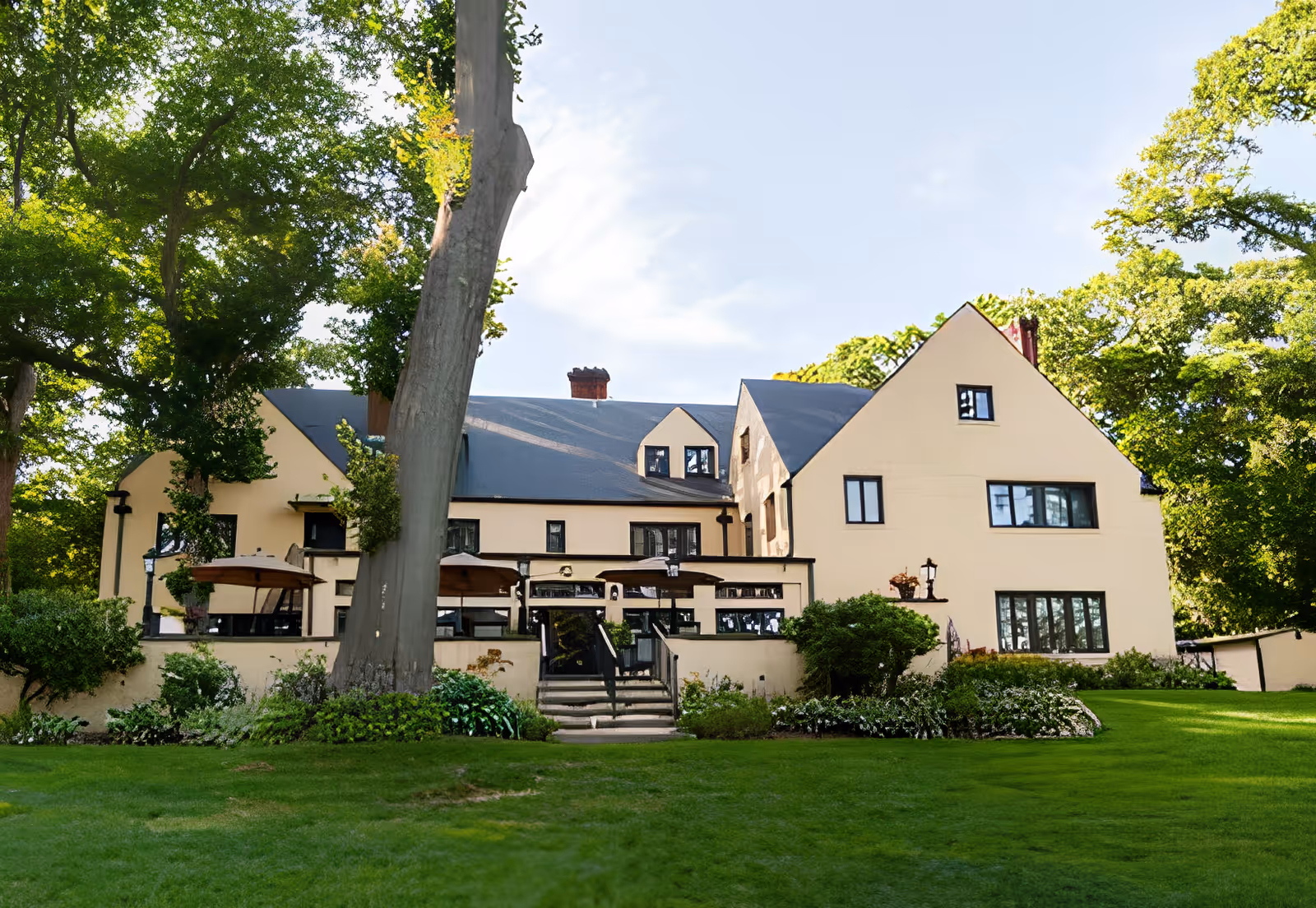 Cream-colored multi-story building with a patio and umbrellas, set behind a manicured lawn and large trees.