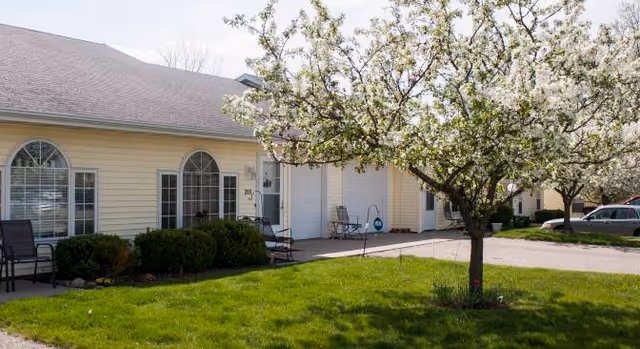 Exterior view of a single-story senior living facility building with yellow siding, arched windows, and a blossoming tree in the foreground on a grassy lawn.