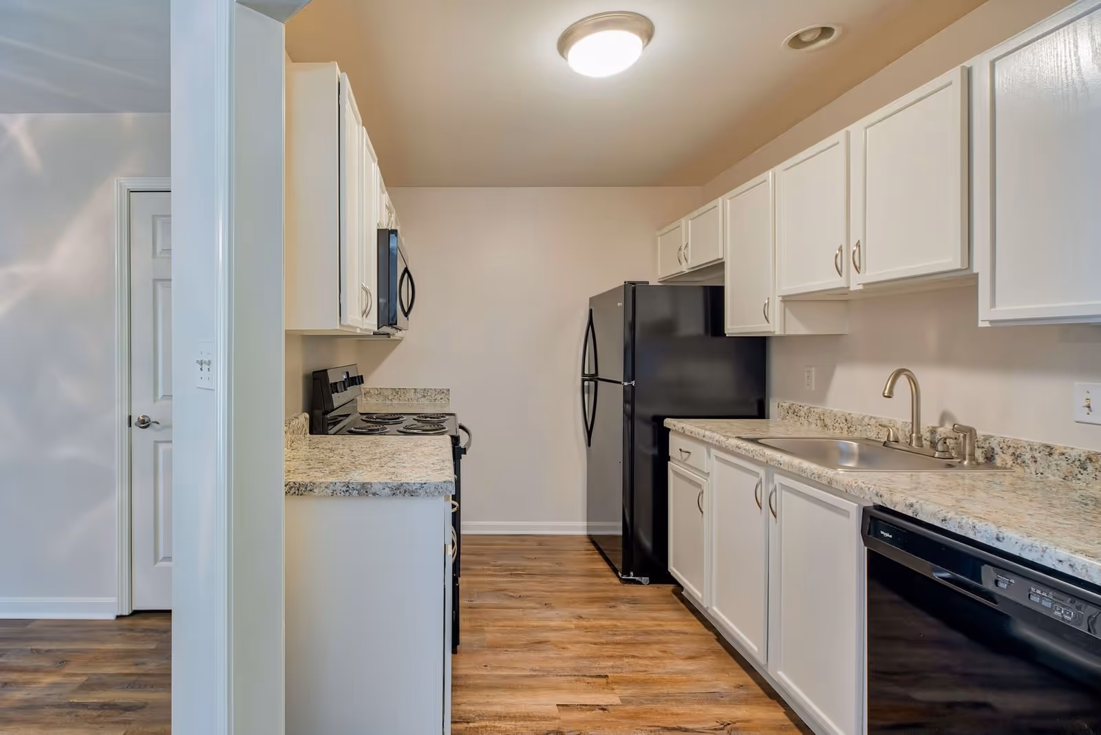 A modern kitchen with white cabinets, granite countertops, a black refrigerator, black dishwasher, black stove with oven, and a stainless steel sink. The floor is wood-style laminate, and the walls are painted beige. The kitchen is well-lit with a ceiling light.