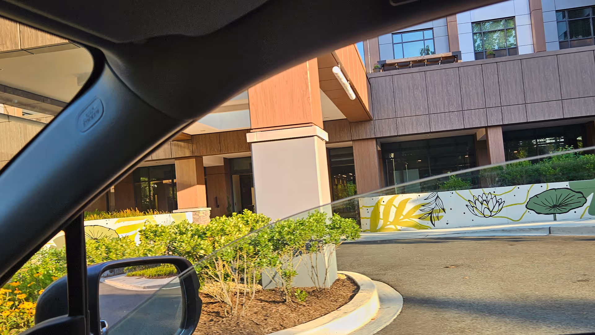 View from a car of a modern senior living facility exterior with landscaped entrance and mural.