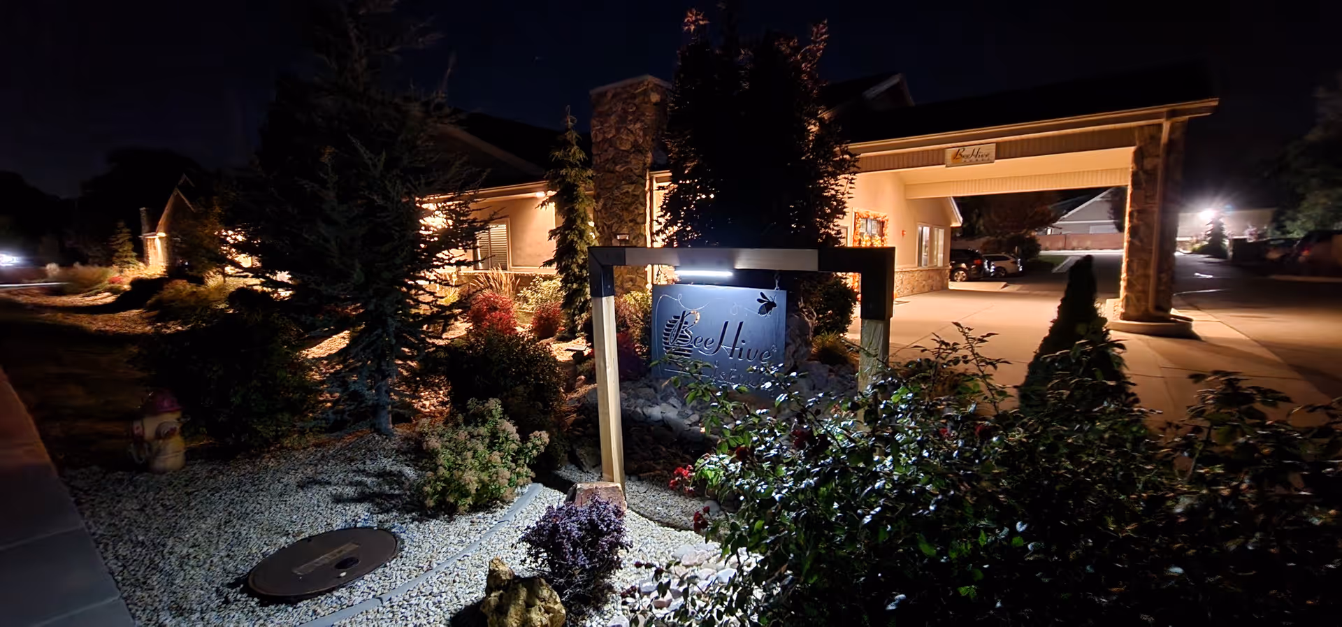 Night view of the BeeHive Homes entrance sign and landscaped front driveway illuminated by exterior lights.