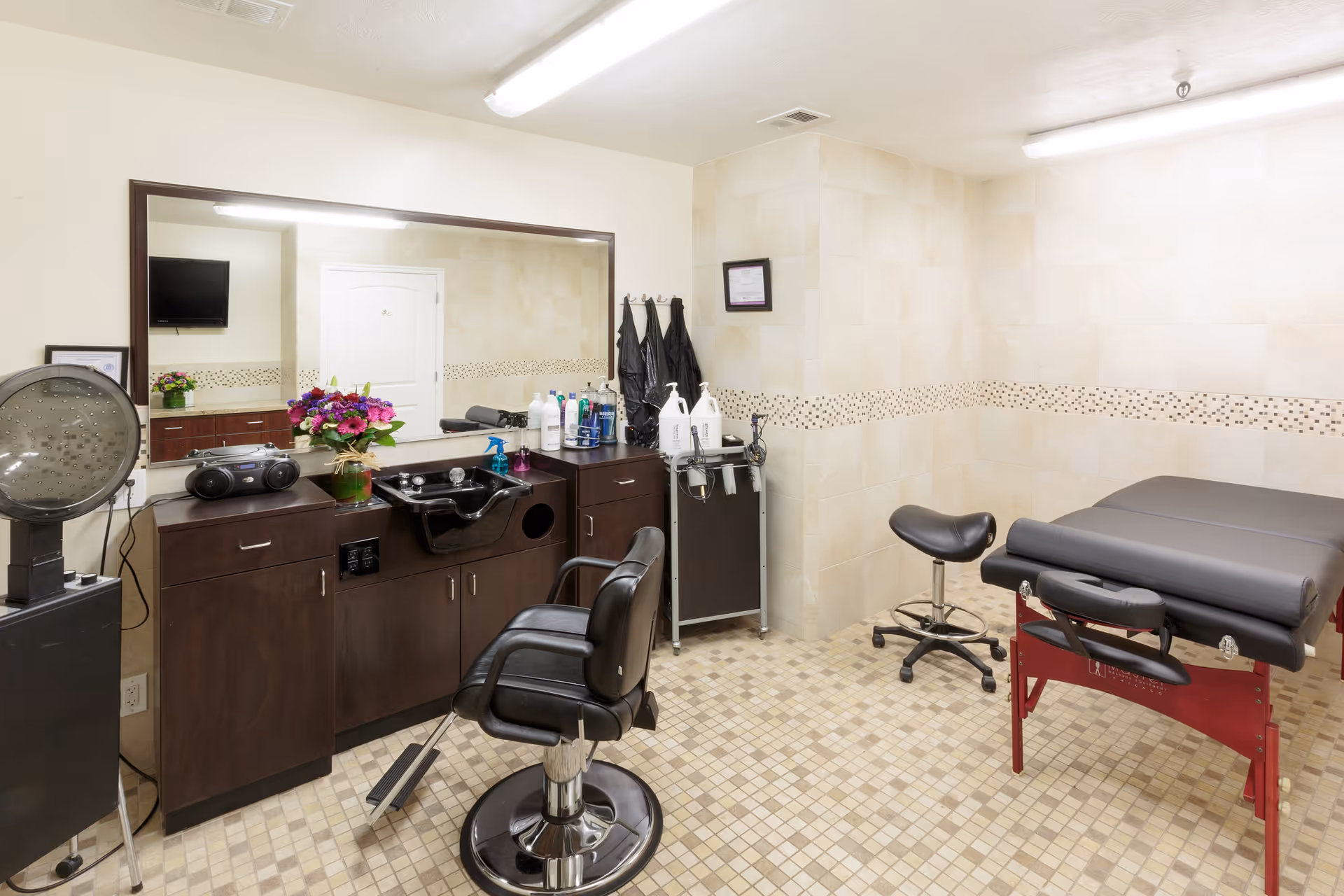 A well-lit room with a large mirror above a dark wooden cabinet holding hair care products, a black salon chair, a hair dryer, and a black massage table with a matching stool on a tiled floor.