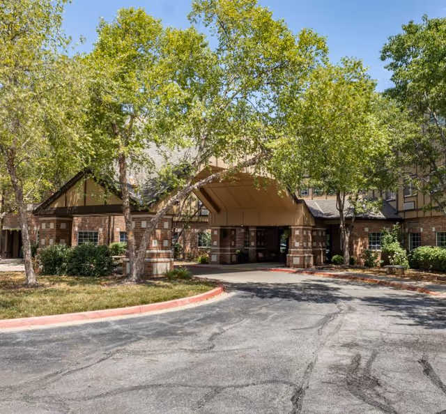 Brick senior living facility with a covered entrance and green trees along a curved driveway