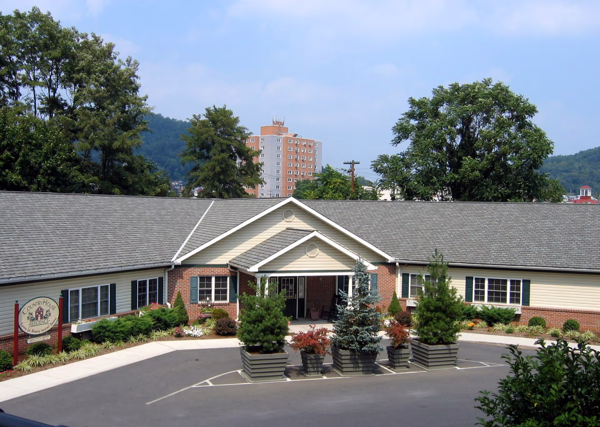 Exterior view of CountryHouse Residence, a single-story building with a gray shingled roof, brick and beige siding, surrounded by landscaped greenery and potted plants in front of the entrance. Trees and a tall apartment building are visible in the background under a partly cloudy sky.