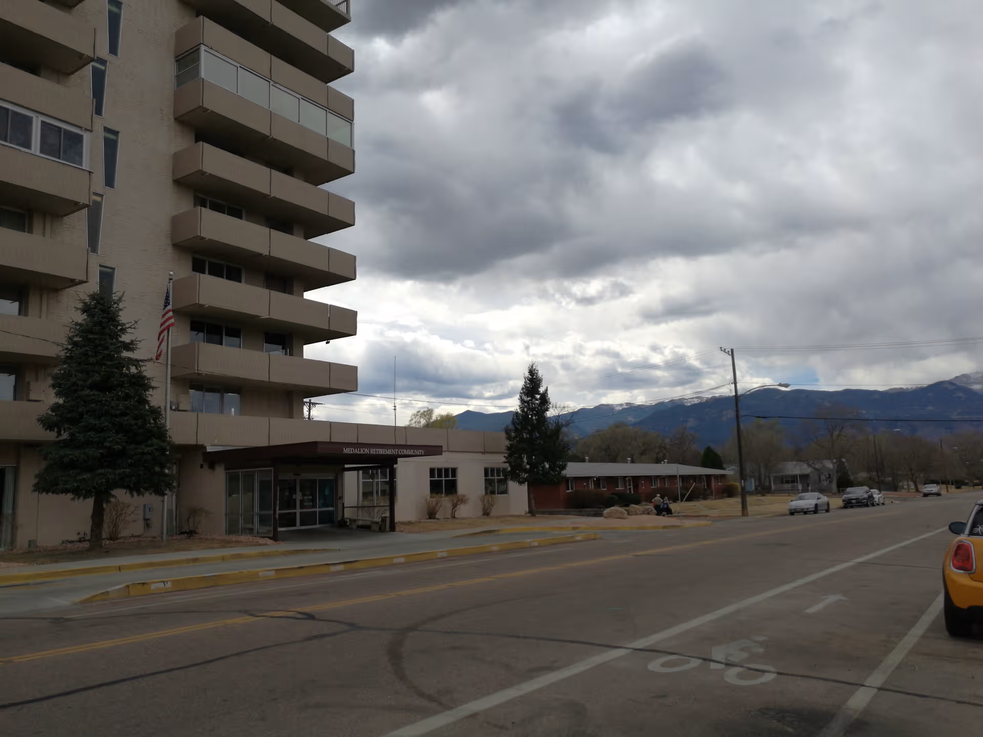 Front view of a tall beige building with a covered entrance and distant mountains.