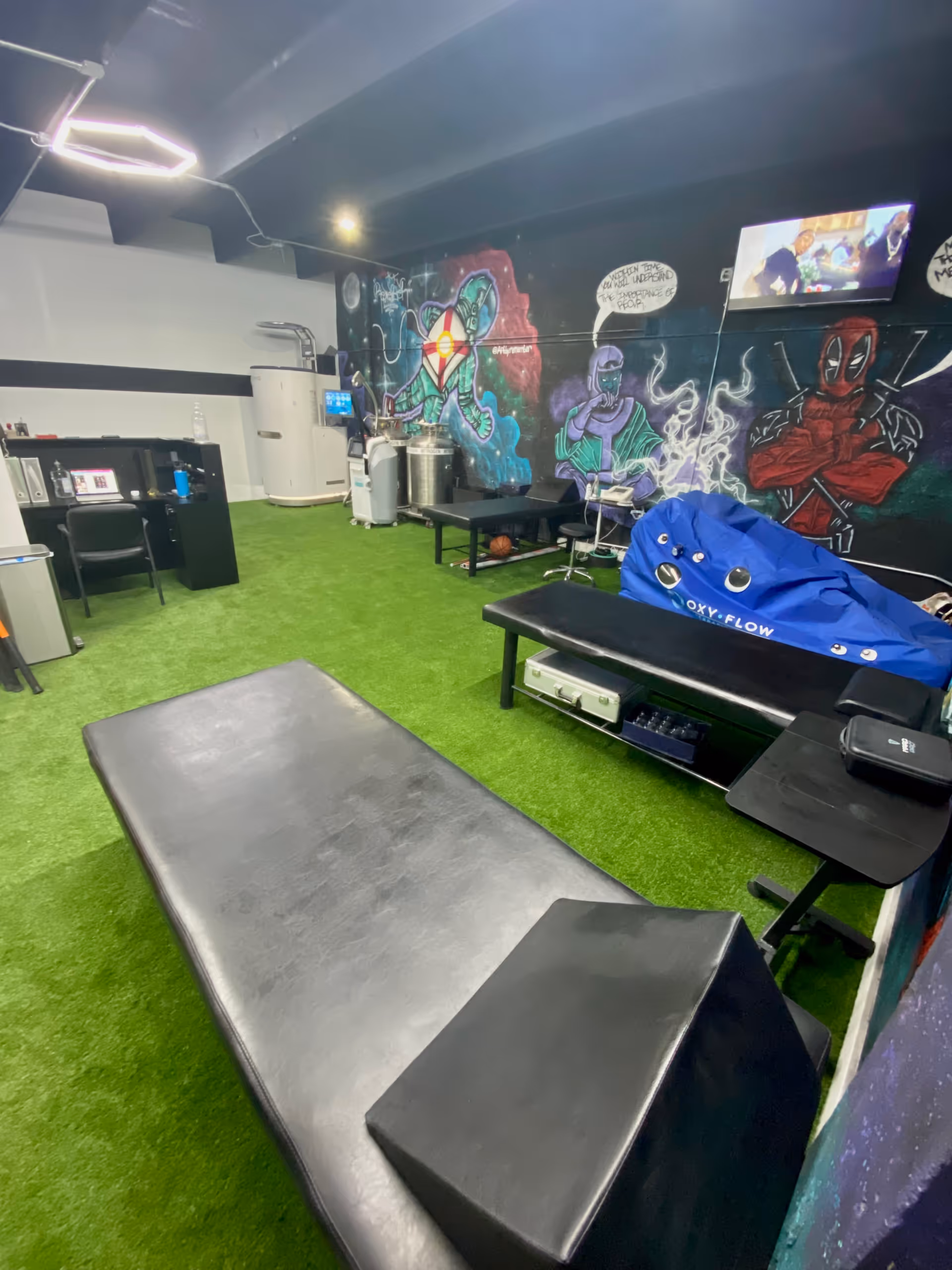 Indoor rehab gym with turf flooring, therapy tables, graffiti art wall, and equipment.