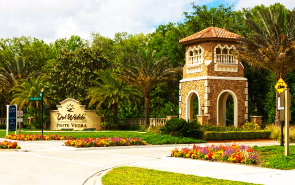 Entrance to Del Webb Ponte Vedra featuring a stone tower structure with arches and a tiled roof, surrounded by palm trees and colorful flower beds under a partly cloudy sky.