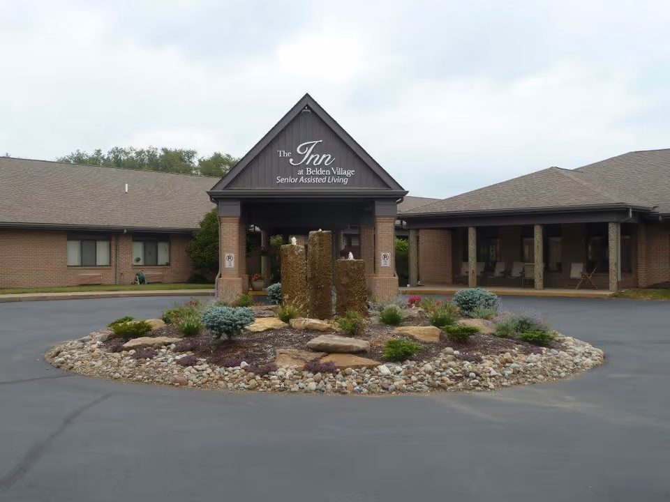 Front exterior view of The Inn at Belden Village Senior Assisted Living facility showing a circular driveway with a landscaped rock garden and water feature in the center, and a covered entrance with brick pillars.
