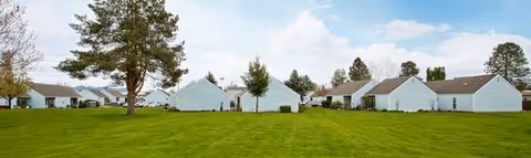 Wide green lawn in front of white, single-story buildings and scattered trees.