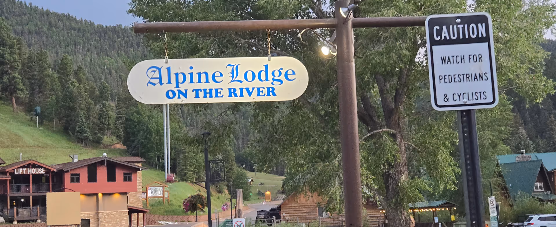 Outdoor scene with Alpine Lodge on the River sign and caution sign beside rustic buildings.